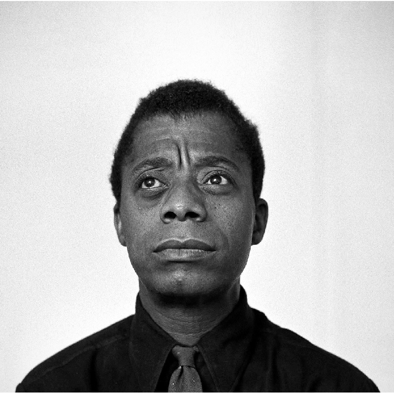 Foto em preto e branco de James Baldwin sobre fundo branco. Ele olha alto para o horizonte.