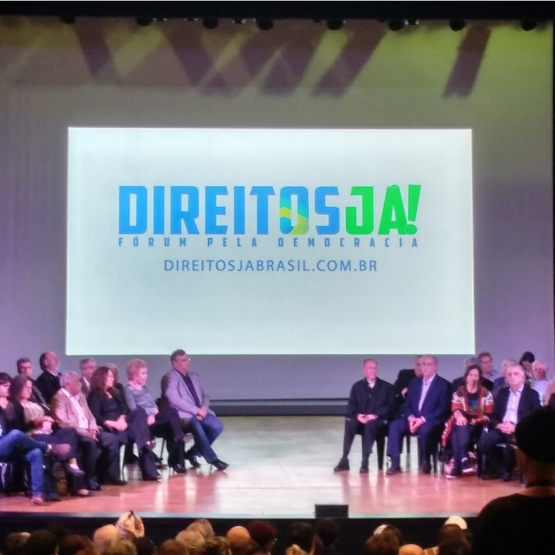 Palco do TUCA no dia do evento "Direitos Já". Uma projeção com o slogan da campanha ao fundo e diversas lideranças políticas sentadas à frente.