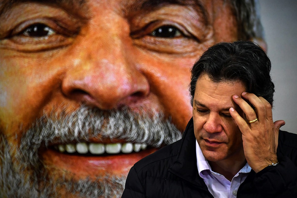 A imagem mostra Fernando Haddad cabisbaixo e um Lula atrás sorridente.